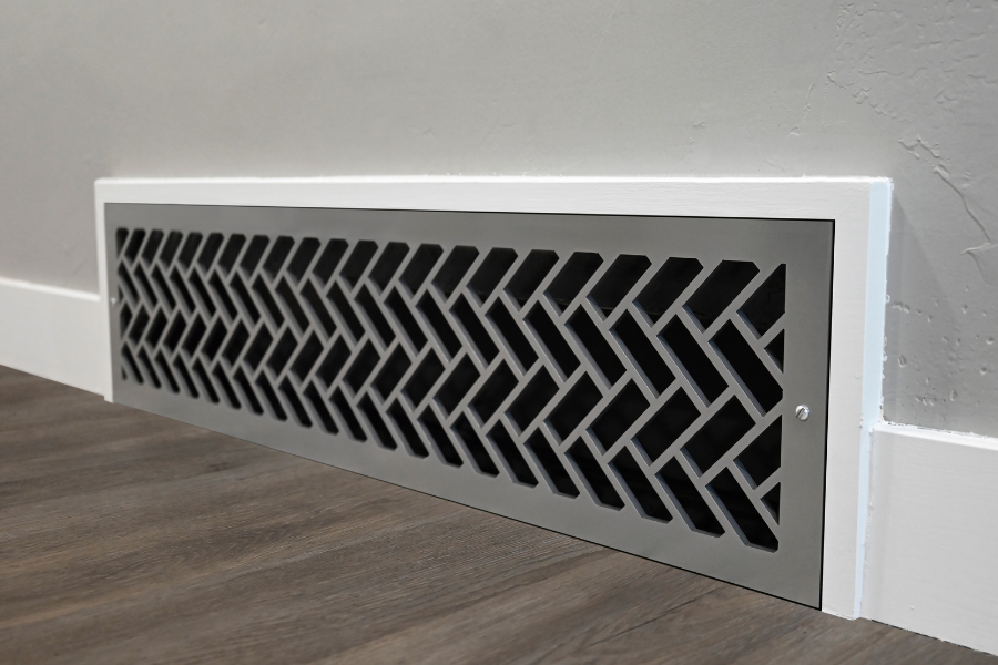 A rectangular decorative metal air return vent with a geometric chevron pattern, installed against a white wall on a wood-look floor.