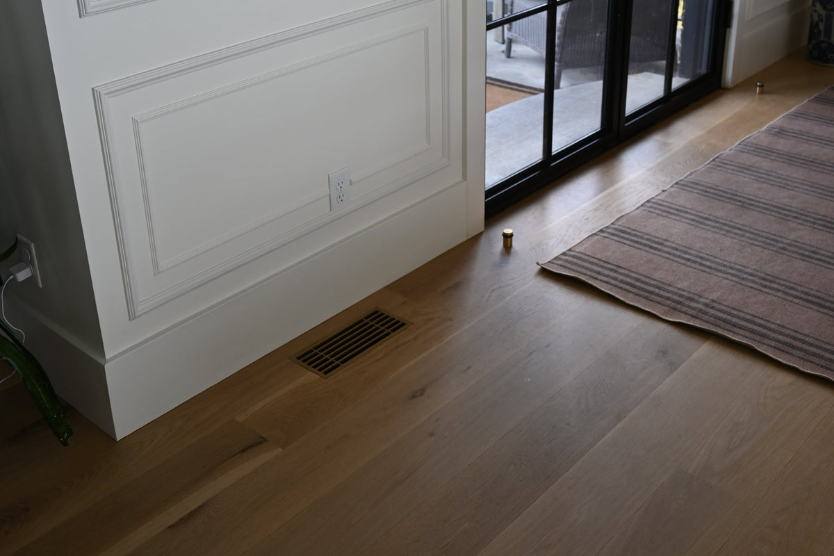 Light brown hardwood flooring installed in a modern room with white paneled walls, a black-framed glass door, and a striped area rug next to a decorative metal vent with modern lines installed flush into the wood flooring.