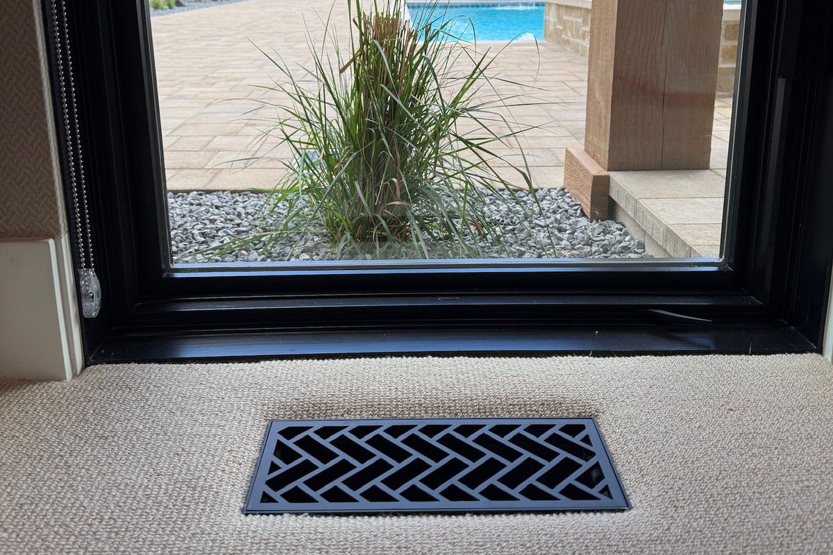 A black flush mount decorative metal floor vent in carpet with a geometric herringbone pattern, installed on a beige carpeted floor in front of a window looking out onto a patio and swimming pool.