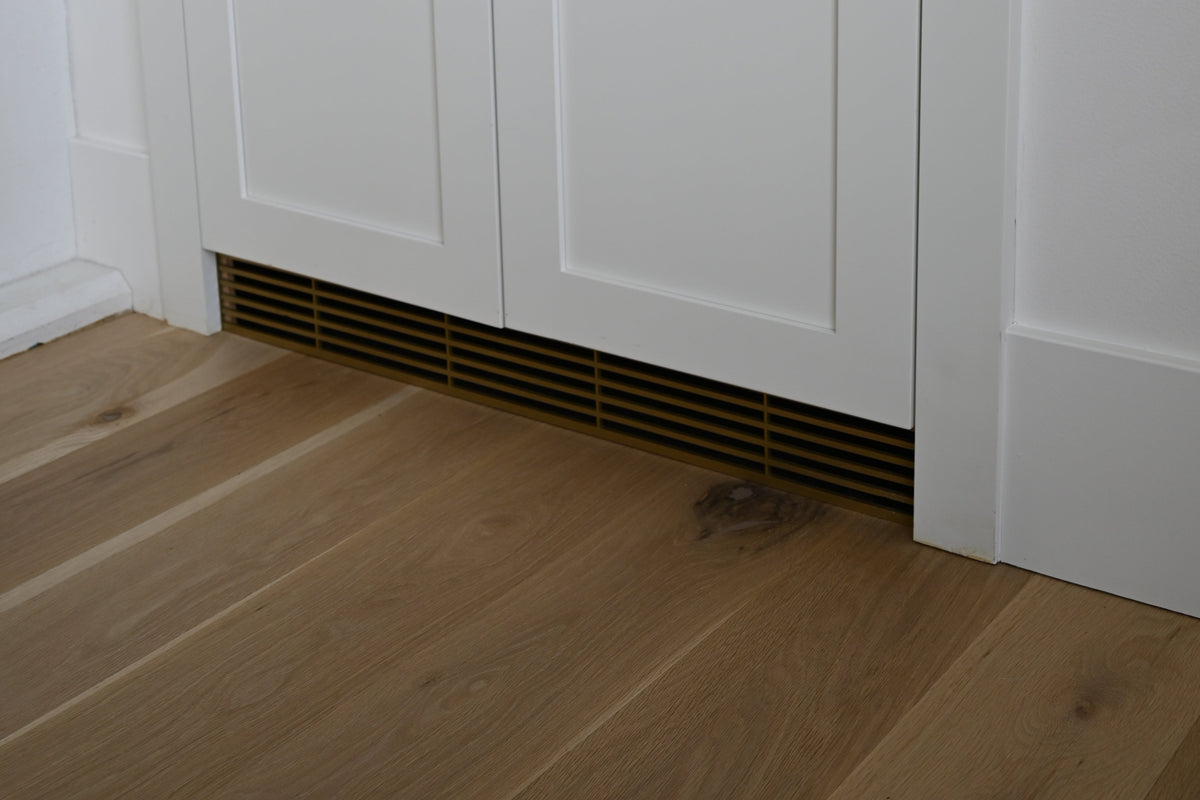 Custom modern toe kick made out of metal in a White built-in cabinet with two paneled doors and small round gold knobs, installed in a white wall with light wood flooring and a patterned storage box on top.