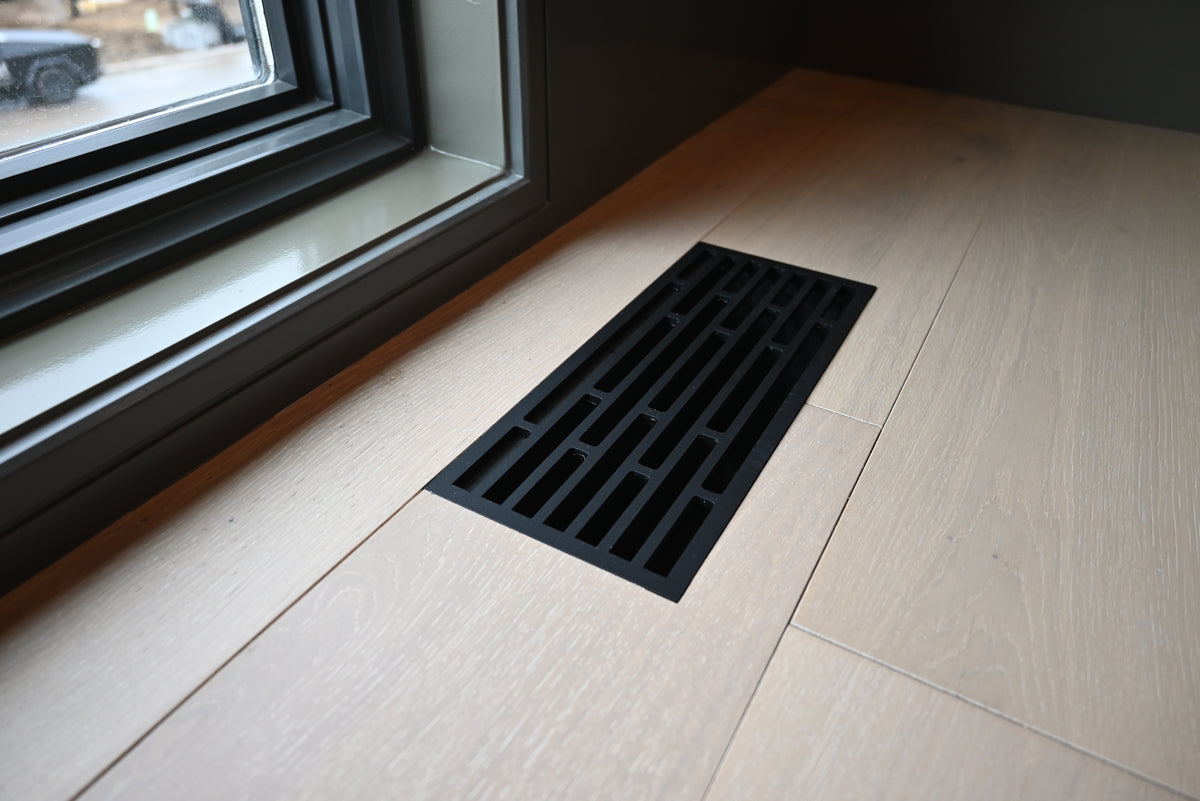 Flush Mount Vent - Straight - Matte Black. A rectangular black metal floor vent grate installed on a light beige wood-look tile floor next to a dark-framed window.