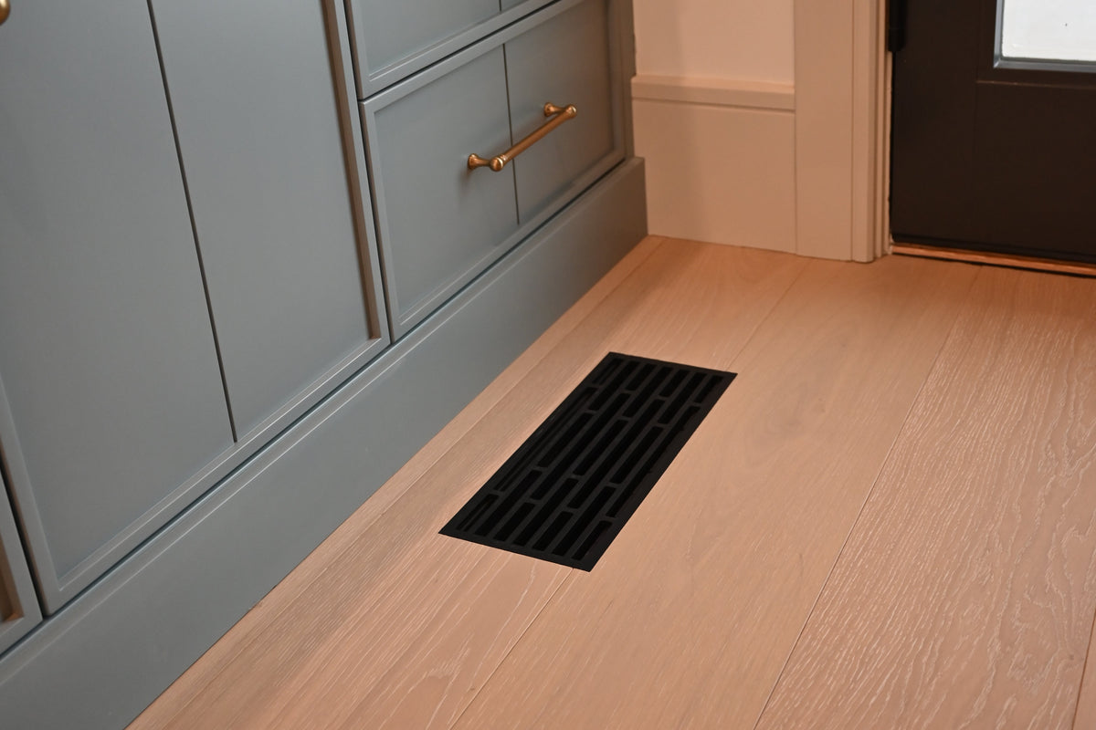 A light gray kitchen cabinet with brass handles and a marble-look top, shown in a real interior space with light wood flooring and a black flush mount floor vent.