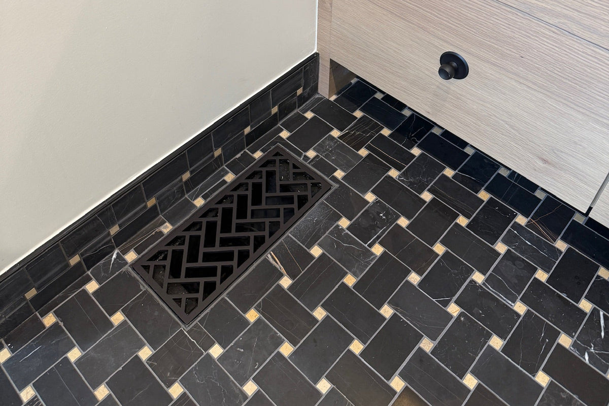 A close-up view of a bathroom corner showing a black and white herringbone tile floor with a decorative metal flush mount vent installed into tile, a light wood vanity cabinet with black knobs, and a beige wall.