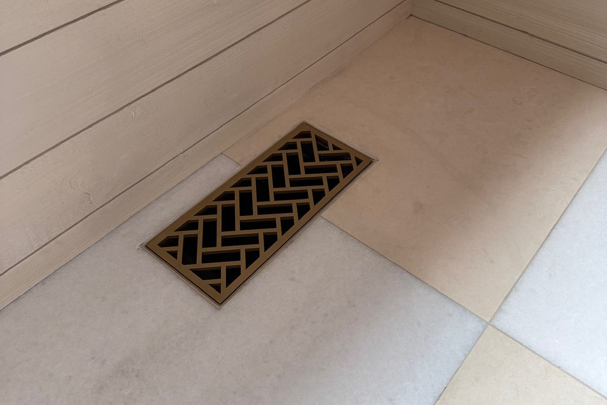 A rectangular flush mount tile floor vent with a black and bronze geometric chevron pattern, installed in the corner of a light beige tiled shower or bathroom floor against wood-paneled walls.