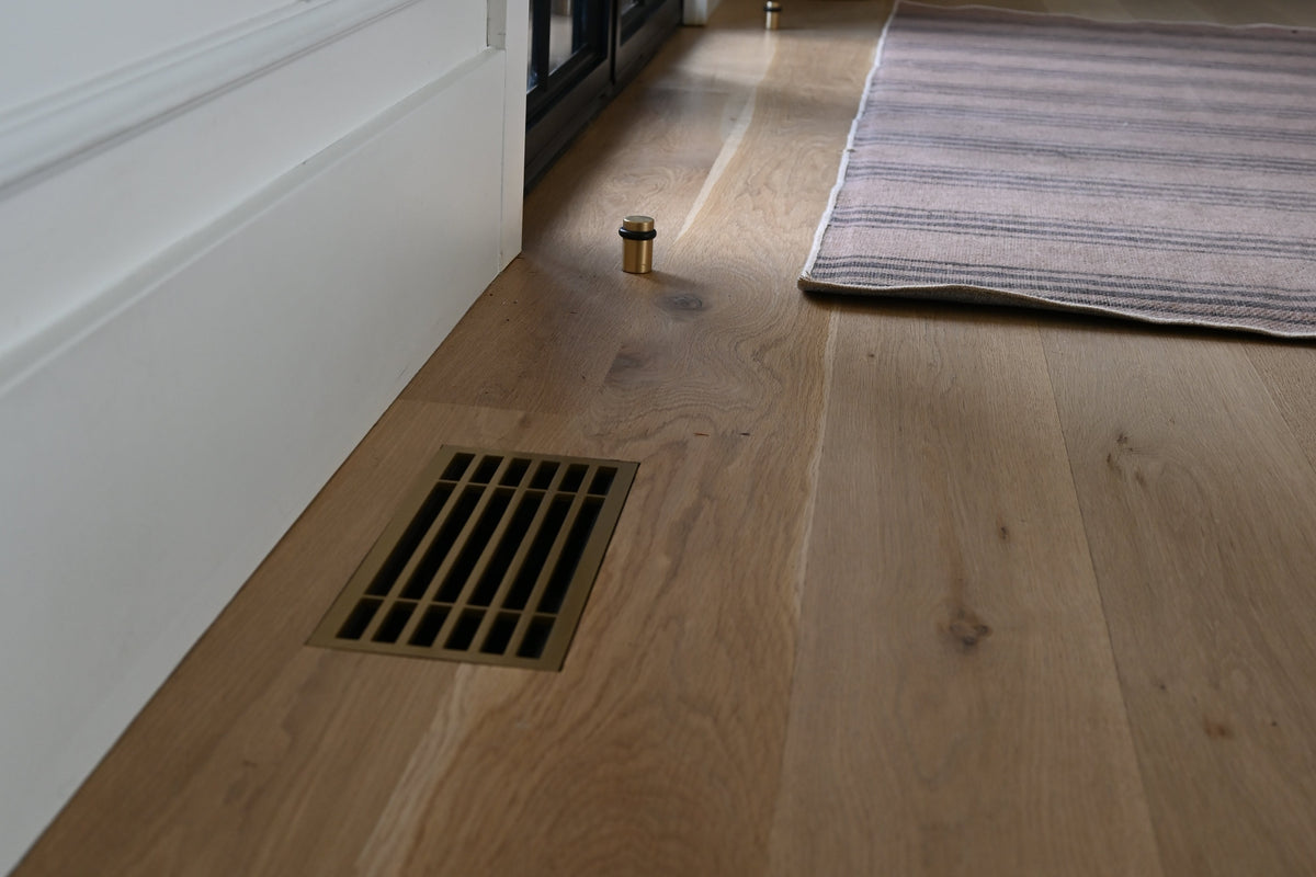 Light oak hardwood flooring installed in a home interior, showing the wood grain, a floor decorative metal vent installed flush into the hardwood floors, white baseboards, a striped rug, and a cabinet in the background.