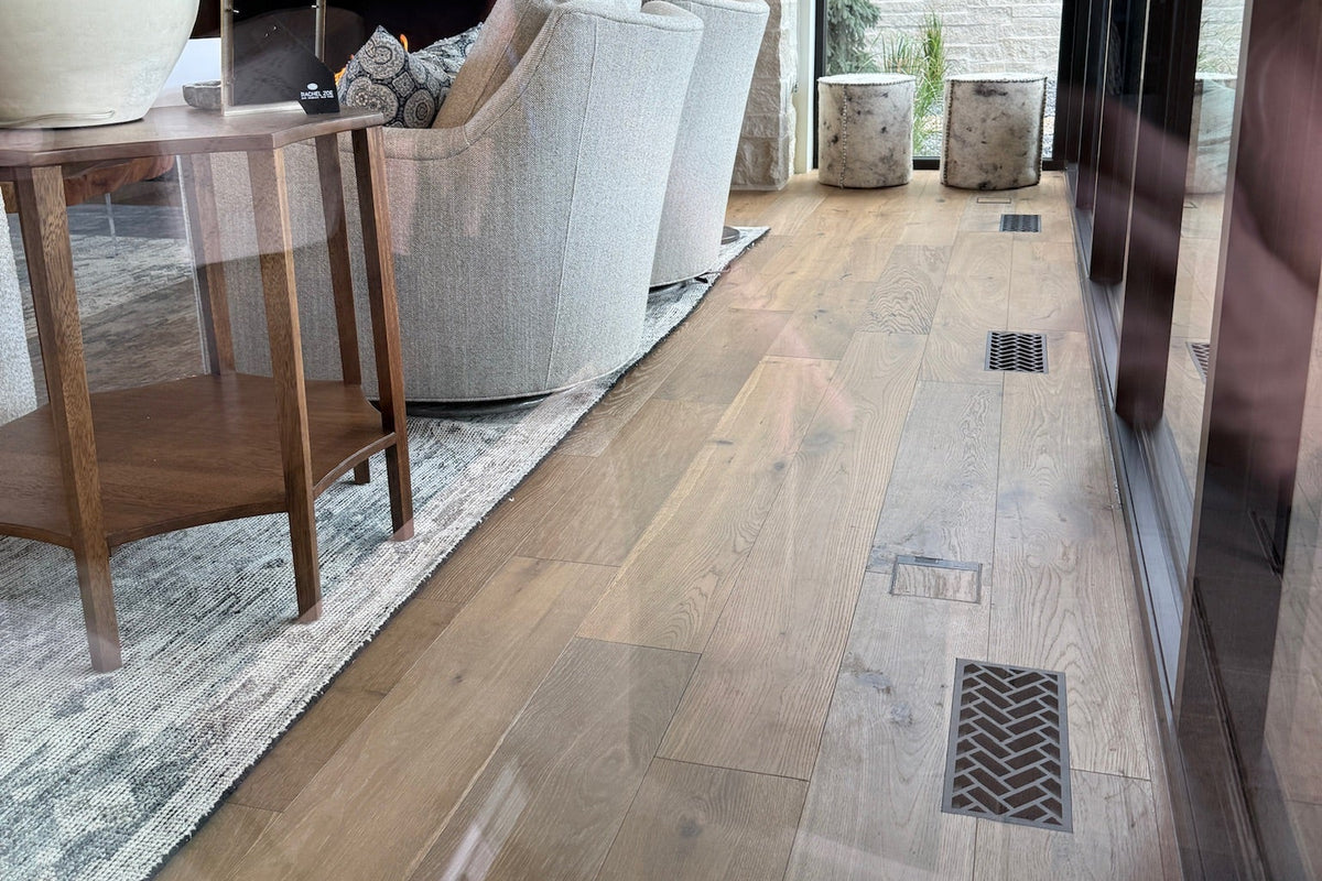 Light oak hardwood flooring installed in a modern living room setting, showing a glossy finish with visible wood grain and flush mounted decorative metal vents with a herringbone pattern.