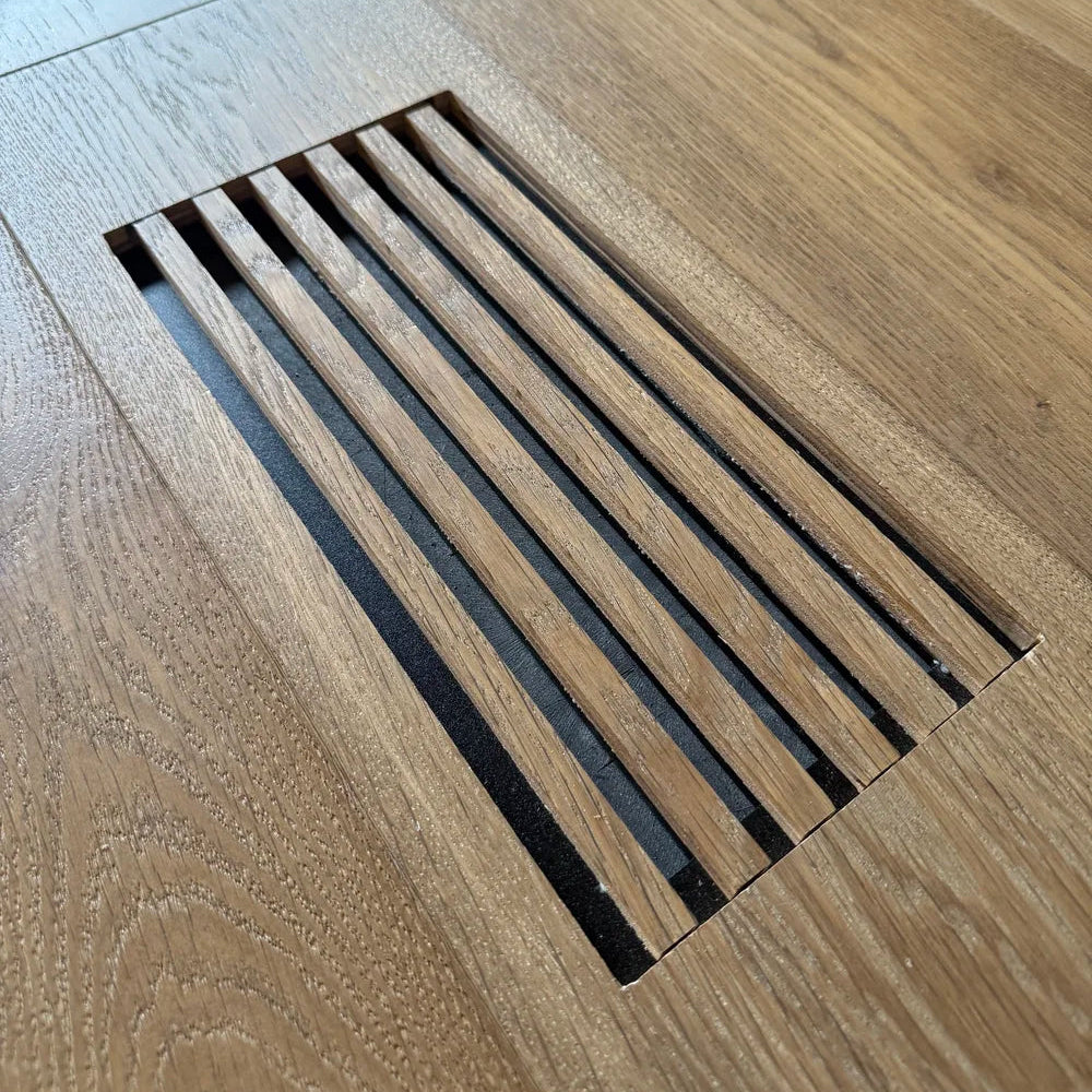 Close-up of a rectangular floor vent with parallel slats set into light oak wood flooring.