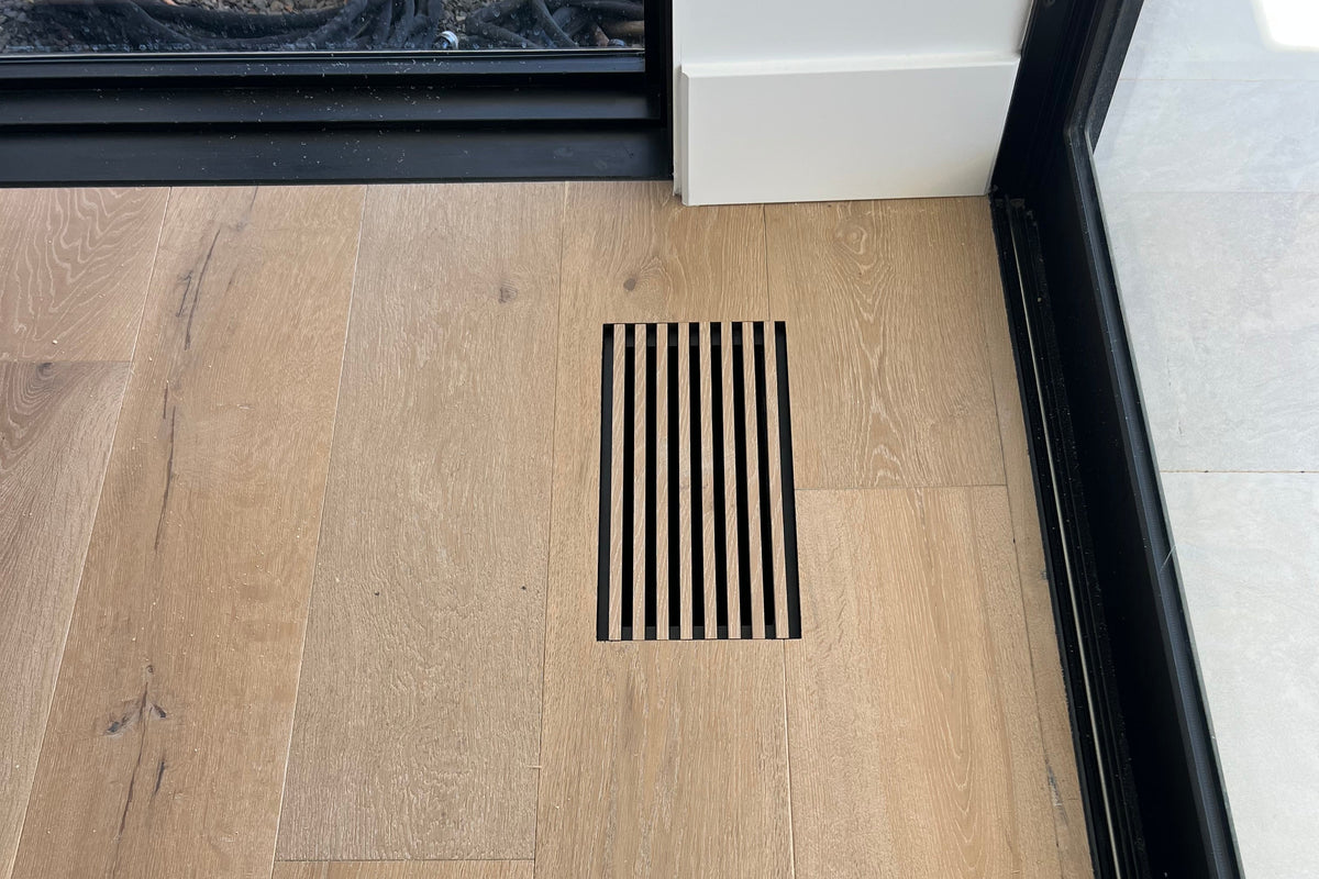 A rectangular floor vent made from flooring material installed on light oak wood flooring in a room with white walls and sliding glass doors.