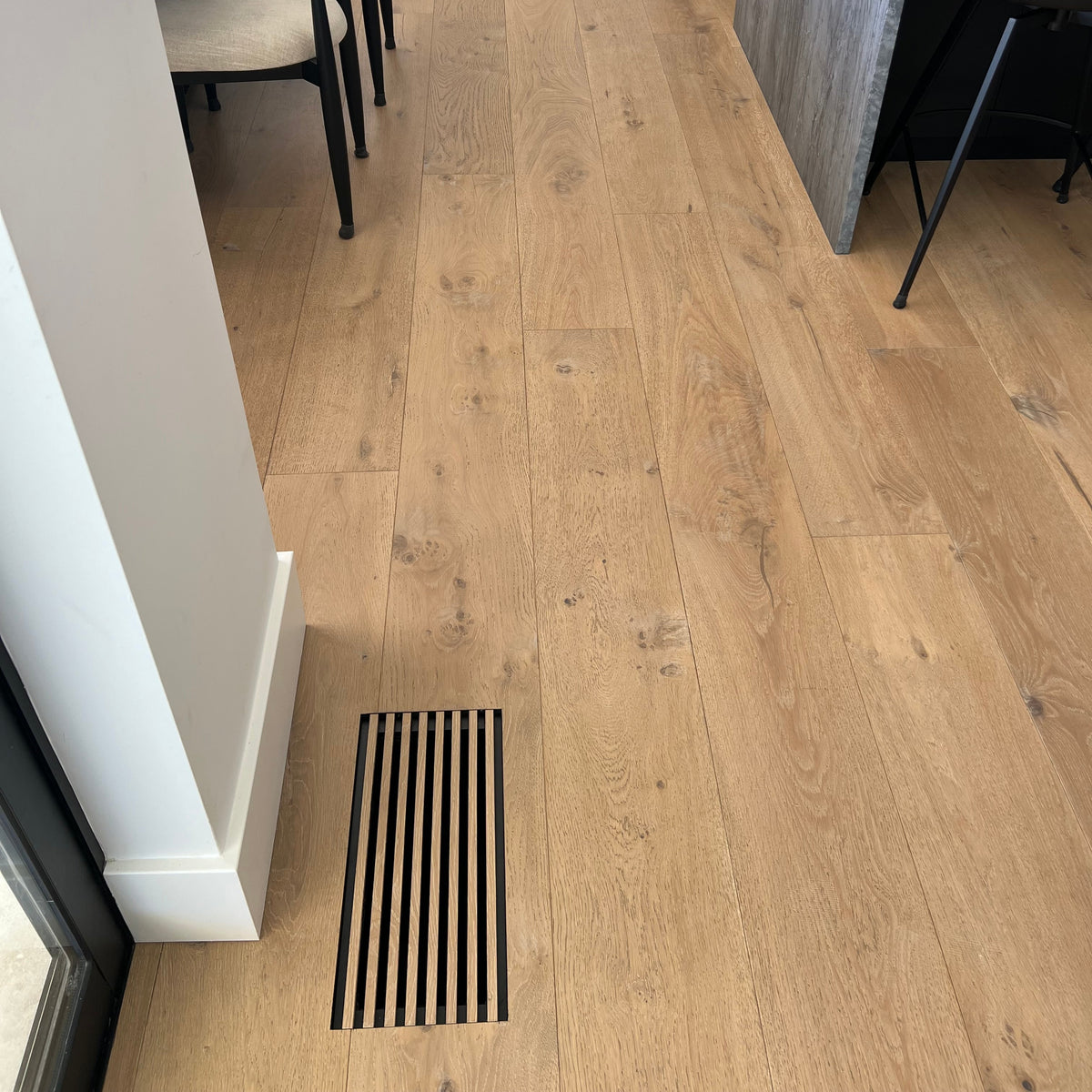 Light oak wood plank flooring installed in a modern kitchen and dining area, showing the floor texture and a floor vent made out of flooring material near the entrance.