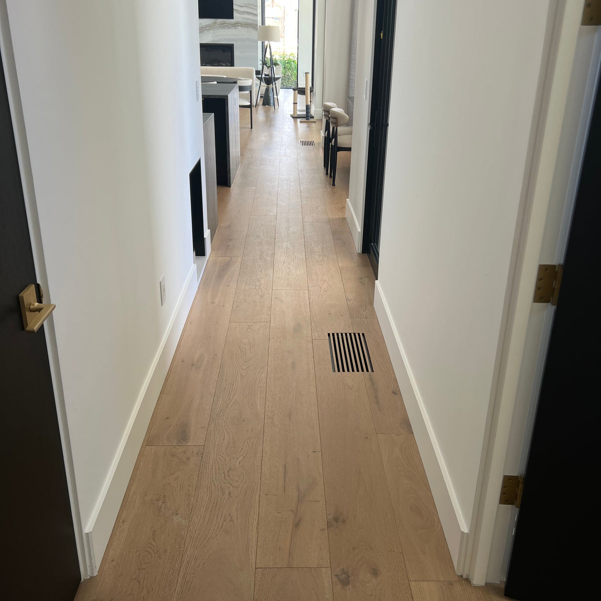 A long hallway with light oak wood plank flooring, white walls, and dark doors on both sides, leading to a bright room in the distance and a floor vent created from flooring material made with a support plate. 