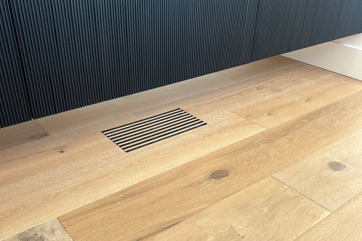 Light oak wood plank flooring shown in a modern interior setting with a ribbed dark gray cabinet and white oak floor vent created by the same material as the floor.