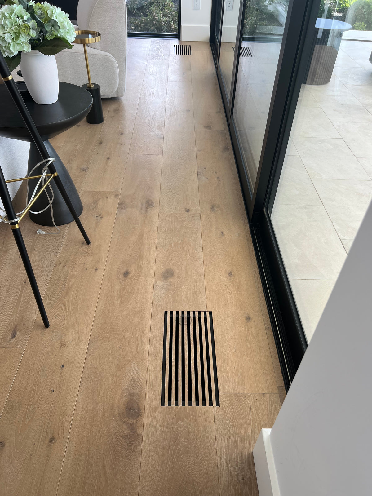 Light oak wood flooring with a slatted floor vent created from the flooring material near the center, shown in a modern home interior next to black-framed glass doors.