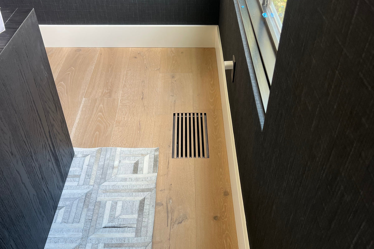 A high-angle view of a modern interior corner showing light oak wood flooring, dark textured walls, a window, and a gray geometric patterned rug partially visible on the left with a white oak floor vent created by the flooring material.