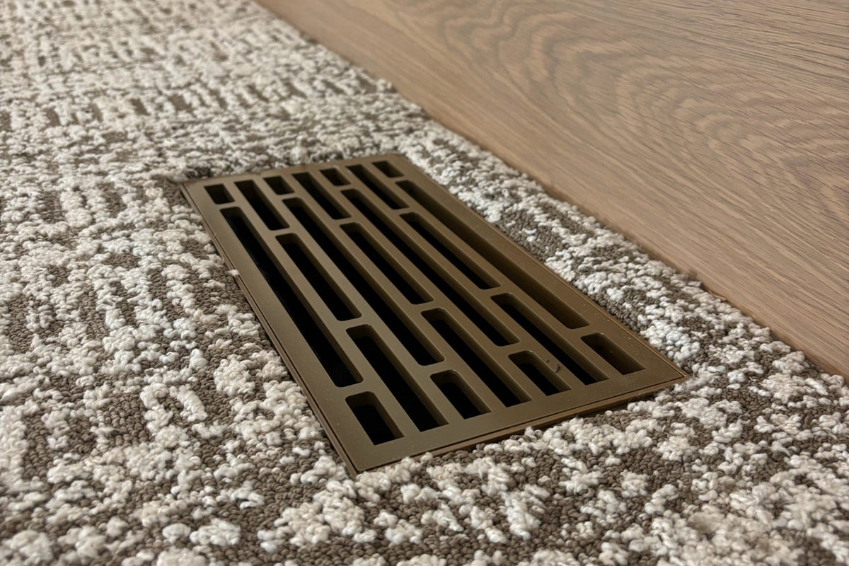 A rectangular metal floor vent with a slotted grate, installed on a textured brown and beige carpet next to a wood baseboard.