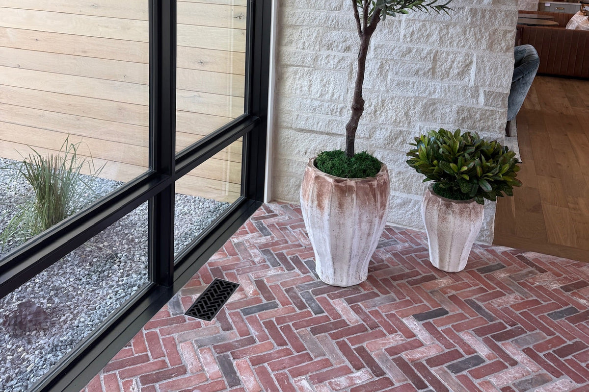 A tall potted olive tree and a smaller green boxwood shrub in textured white planters, set on a red brick herringbone floor with a decorative metal flush mount vent in a herringbone pattern in the floor next to a large black-framed glass window.
